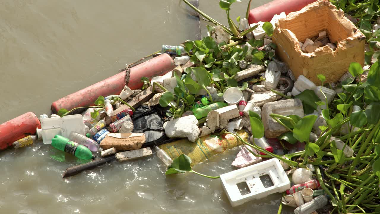 Toma estática en ángulo picado de botellas de plástico y residuos de poliestireno flotando en agua turbia de río.