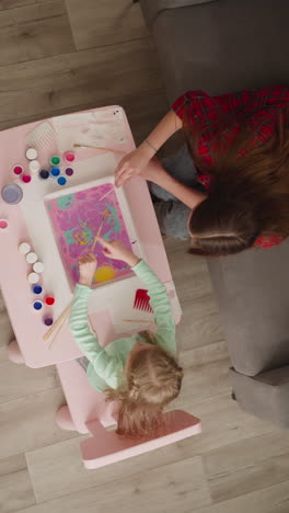 woman and little girl add bright colors to pattern on oily water surface doing ebru drawing in living room upper view. creative hobby for development