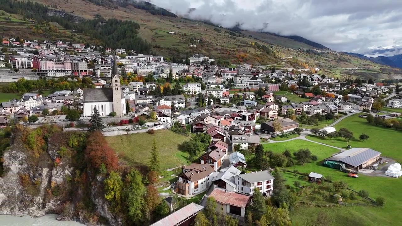 İsviçre'nin Scuol kasabasında antik kiliseli küçük bir İsviçre kasabasının havadan genel çekimi. Yükselen geniş çekim. Evler ve konutlarla bulutlu bir gün.