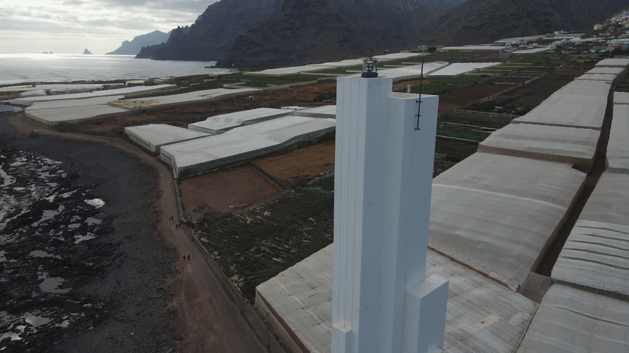 Modern Punta del Hidalgo lighthouse on the rocky coast of Tenerife with greenhouses nearby