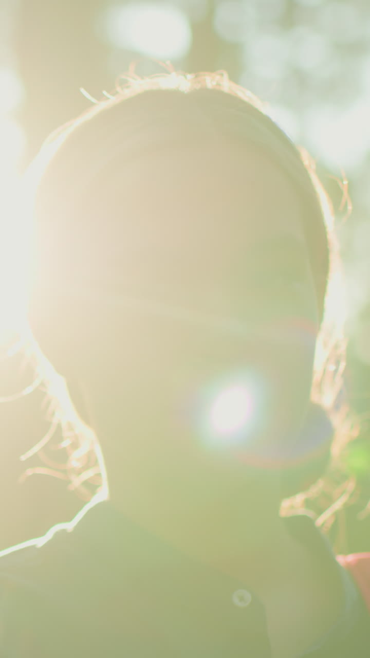 숲 속에 서서 부드러운 미소를 지으며 가방을 조정하는 여인, 빛이 나무를 통해 필터링하는 동안, 빛나는 빛으로 머리카락을 밝히고, 자연 한가운데 따뜻하고 조용한 분위기를 만니다.