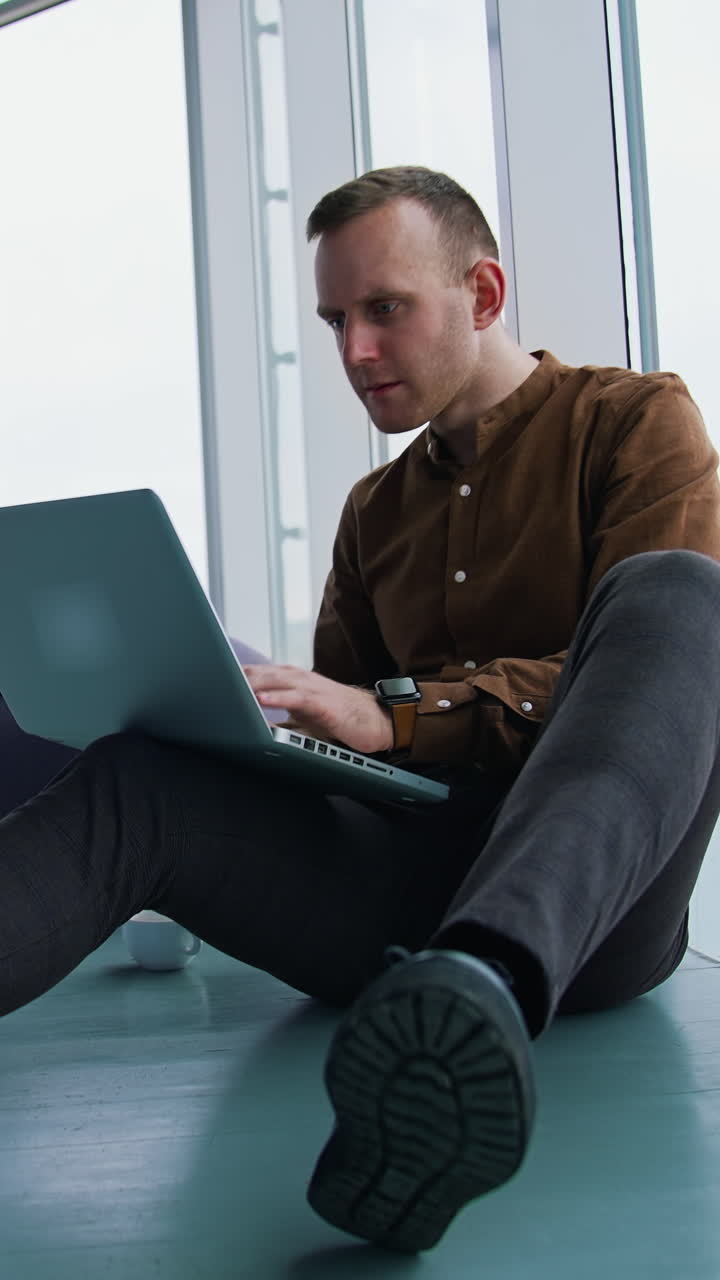 Young man sitting on floor near the window with a laptop. Happy man winning something while using a wireless computer. Vertical video
