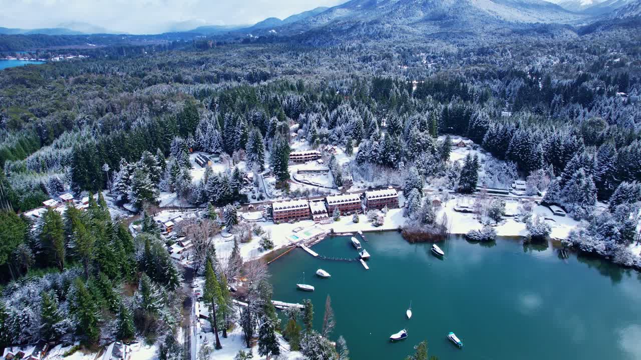 Drone che avanza inclinando la telecamera verso l'alto per rivelare un resort di lusso in riva al lago, barche ormeggiate e le montagne andine innevate sul Lago Nahuel Huapi, Parco Nazionale della Patagonia.