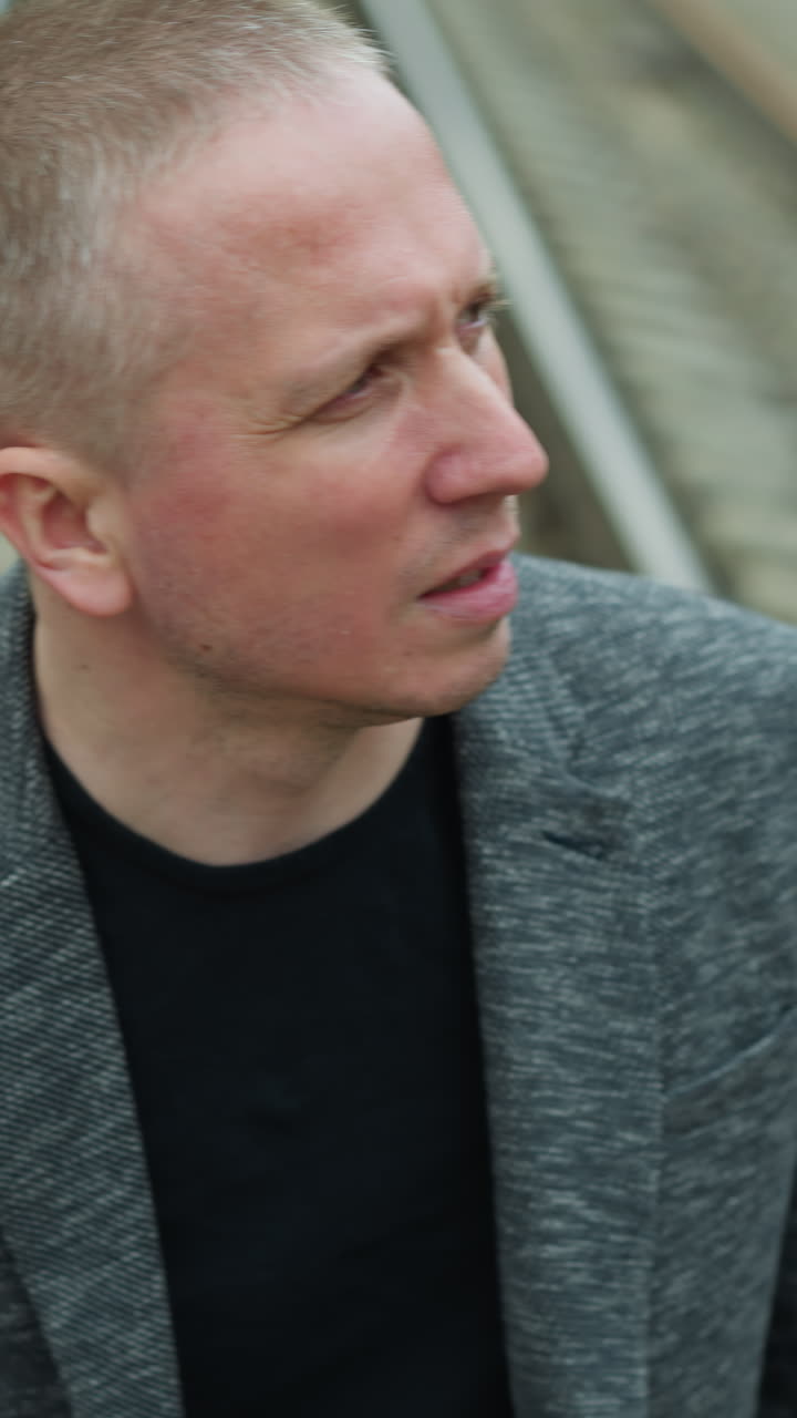 близкий вид пожилого белого человека в сером костюме и черной рубашке поло, отворачивающегося от камеры, идущего по железной дороге с камнями, с размытым видом деревьев и электрических столбов с обеих сторон