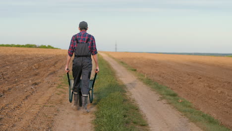 farmer pushing a wheelbarrow on a dirt road through a field