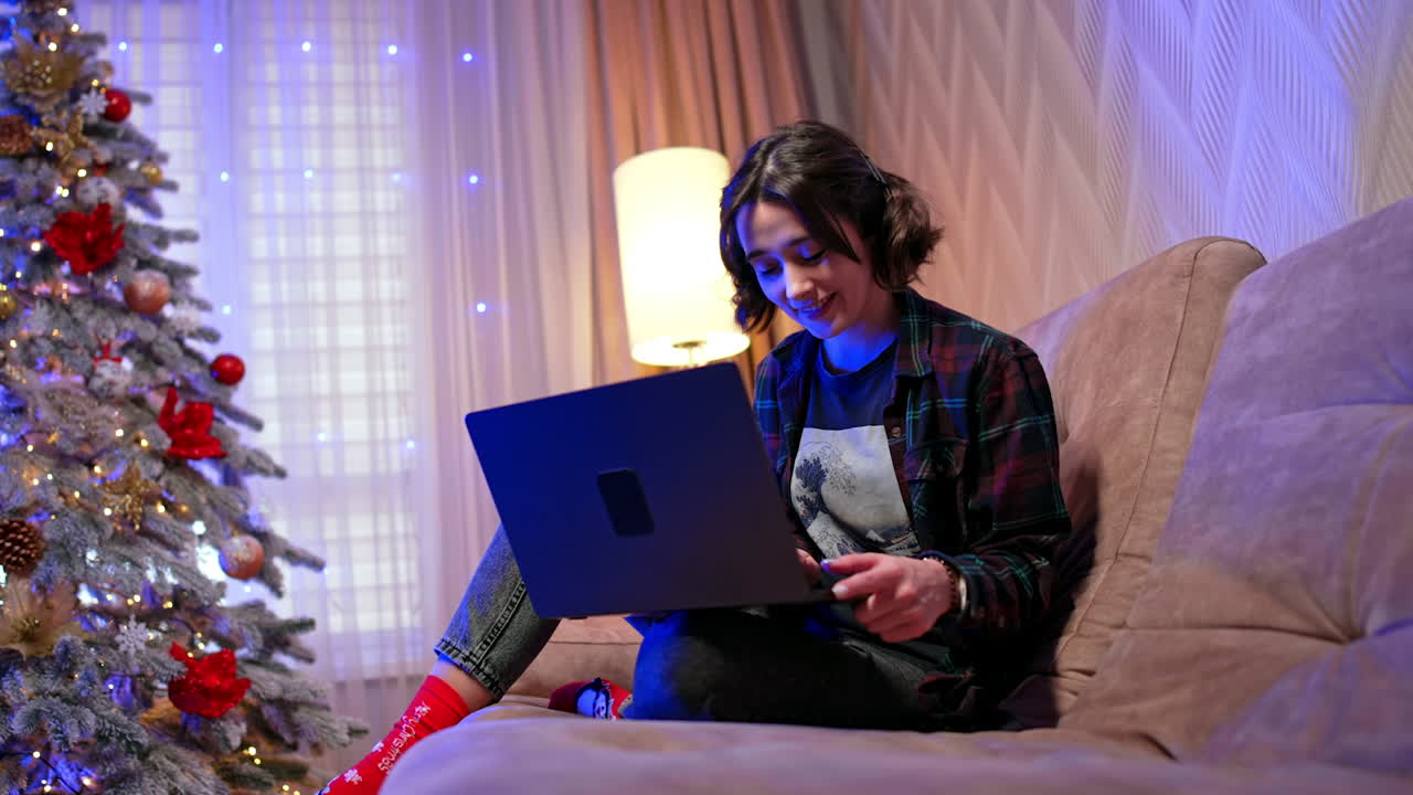 Girl with laptop by tree. A young woman is sitting comfortably on a couch, using her laptop while surrounded by festive Christmas decorations