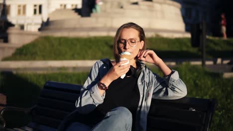 Young-female-in-casual-clothes-enjoying-her-leisure,-sitting-on-a-street-bench-and-licking-big-vanilla-ice-cream.-Delicious