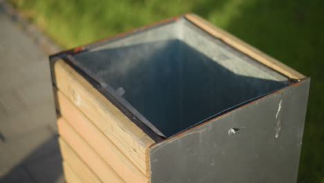 close up of empty wooden and metal waste bin positioned on sidewalk surrounded by green grass in urban outdoor area with soft morning sunlight casting shadows on surface