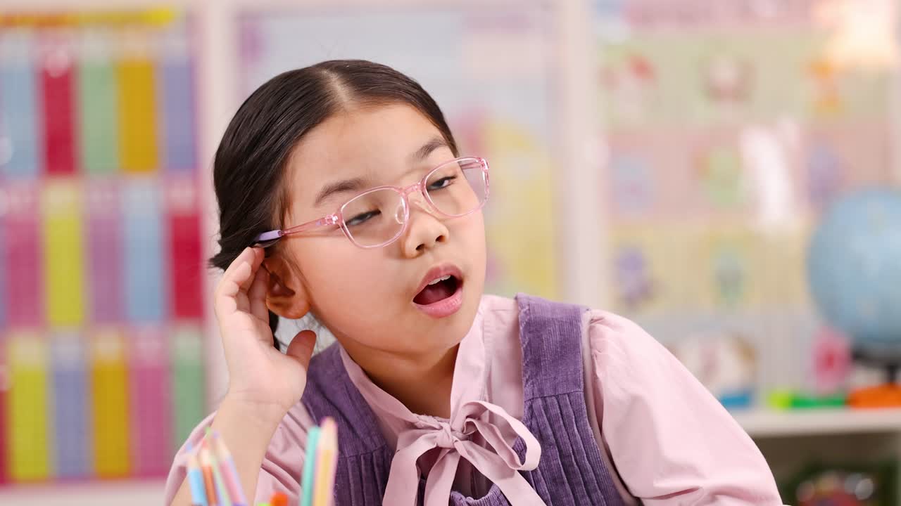 Elementary girl with pink glasses cups ear, listening intently at desk in bright classroom