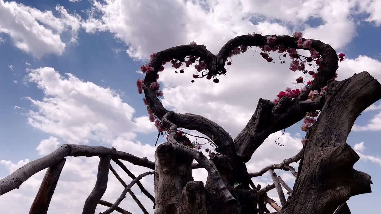 A heart-shaped arbor structure constructed of weathered wood, adorned with delicate pink blossoms, stands against a backdrop of a cloudy blue sky, creating a whimsical and romantic scene