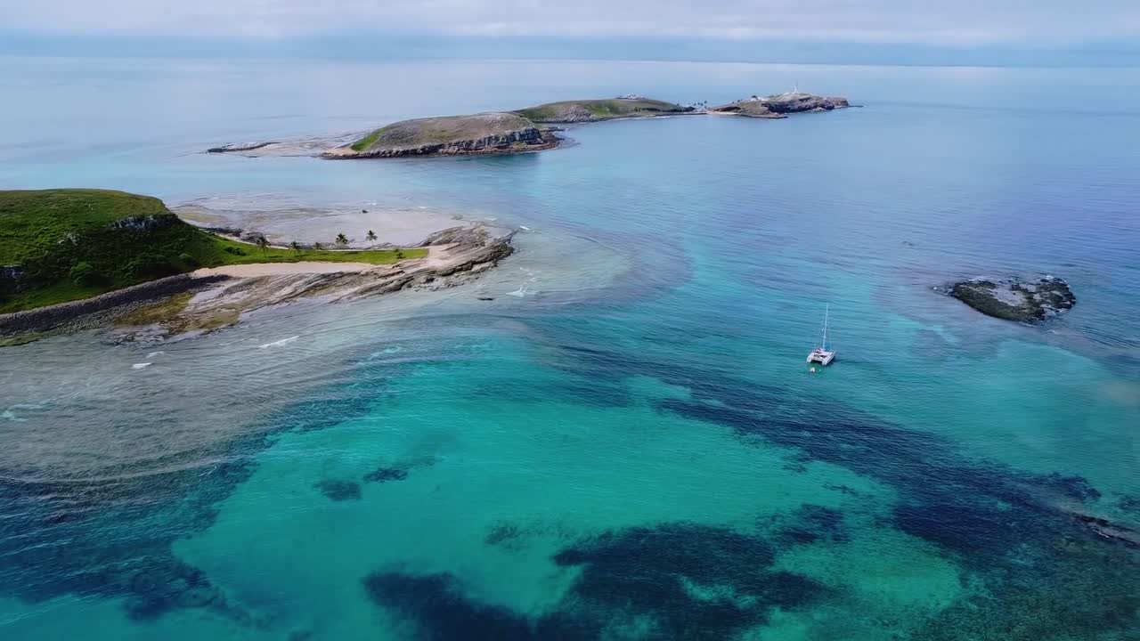 緑豊かな島々に囲まれたサンゴ礁のターコイズブルーの海をカタマランが航行するアブロリョス諸島のパノラマ。豊かな海洋生物多様性と手つかずの熱帯の美しさをドローンパンで捉える。