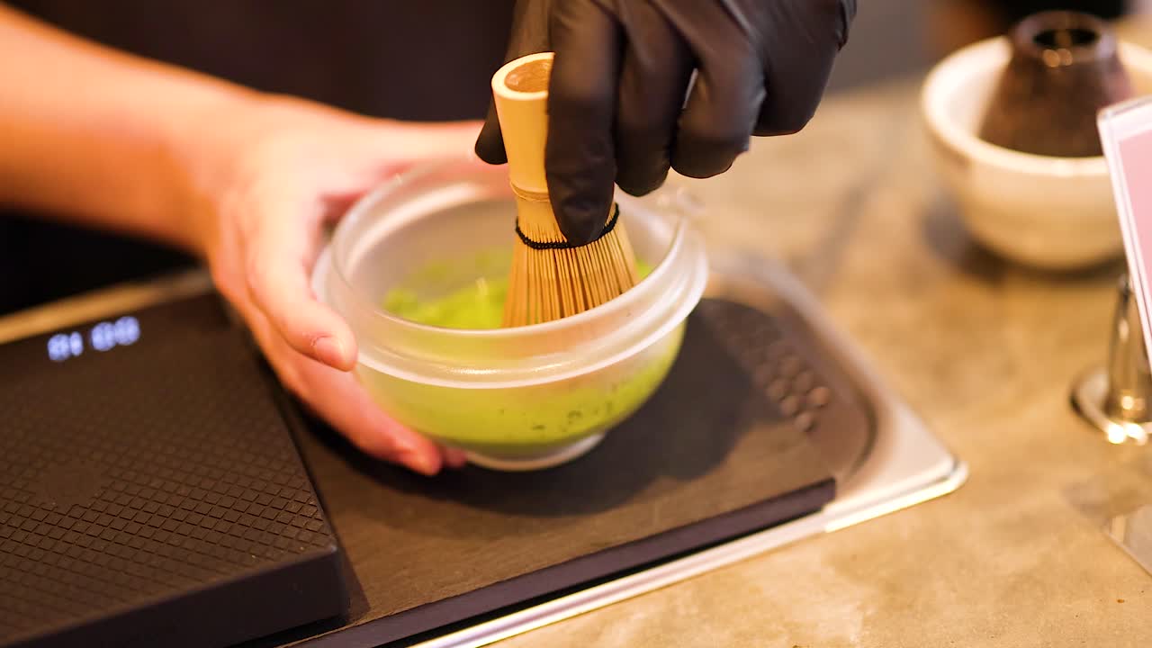 Hands whisk matcha in a bowl using a bamboo whisk. Warm lighting enhances the serene atmosphere of this traditional tea-making process