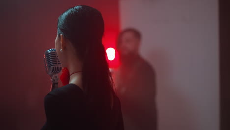 vista trasera de una cantante con una cinta roja en el cuello, cantando en un micrófono vintage con un guitarrista en el fondo rojo borroso