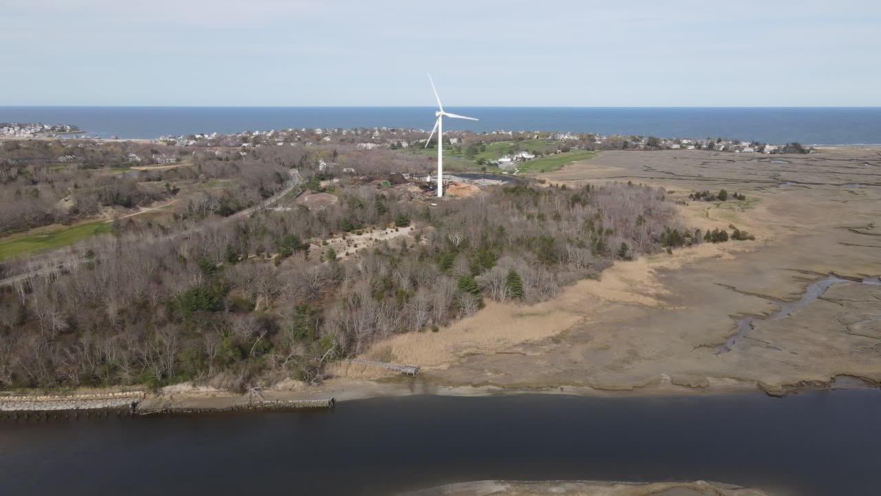 Aerial push over water toward wind turbine on the edge of Scituate Driftway marshes. Atlantic Ocean in the distance.