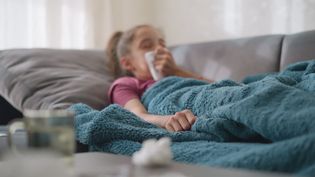 Close up of sick girl sneezing into tissue while lying on couch under warm blanket with glass cup and used tissues on table in foreground indicating cold flu or seasonal illness recovery