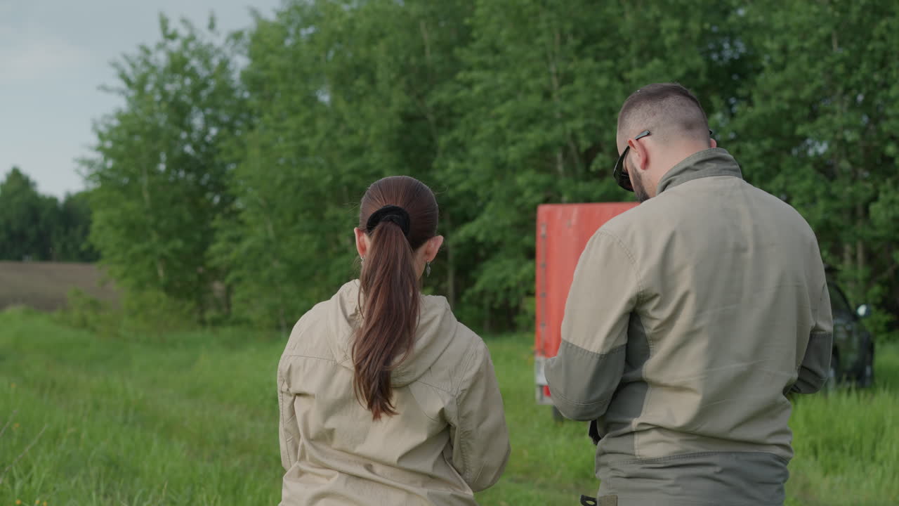 rear view of woman standing beside colleague in green field, both wearing protective jumpsuits and gloves, woman gazing upward thoughtfully while man checks device against lush forest backdrop
