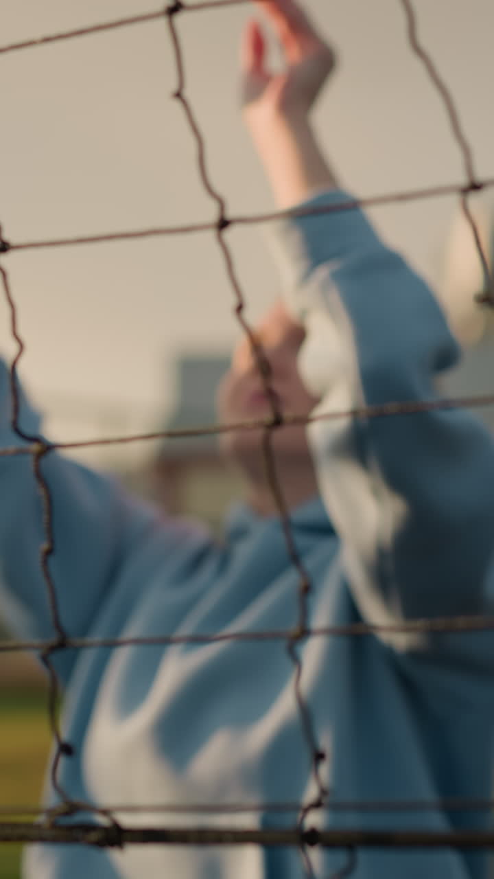 primer plano de la red de voleibol con el jugador visible en el fondo preparándose para un servicio, enfoque suave en la red, mientras que la vegetación y el edificio aparecen borrosos