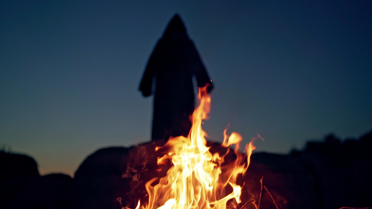 Scary silhouette od black figure on the hill behind fire. Orange flame at night and horrorful death in black cloak with hood is waving arms. Halloween concept.
