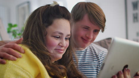 una pareja joven con síndrome de down sentada en el sofá viendo una tableta digital en casa.