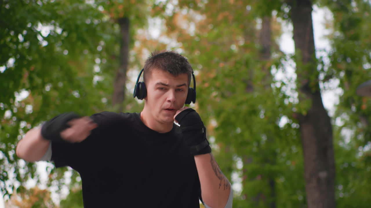 Pria Kaukasia melakukan shadowboxing dengan headphone di luar ruangan, balutan tangan terlihat, ekspresi wajah fokus, latar belakang taman rimbun, kombinasi pukulan sesuai dengan musik, sesi pelatihan energik, nuansa musim gugur