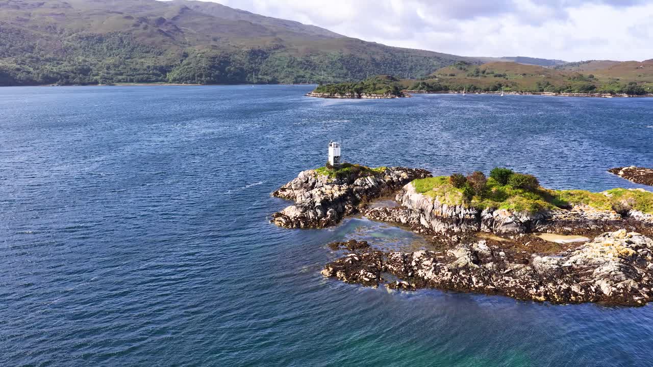 Drone glides over rocky island with lighthouse, blue sea, and distant hills under bright daylight