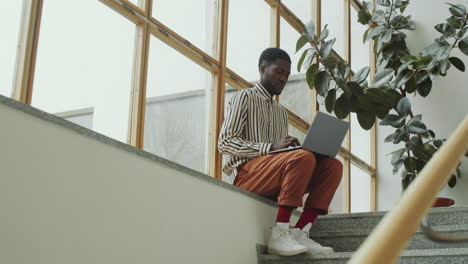 Afro-Man-Sitting-in-Modern-Building-and-Working-on-Laptop