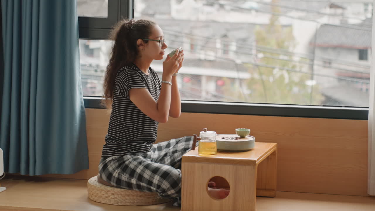 Woman eating hotel breakfast by window, low table with tray and juice, bento and chopsticks on wooden table, sunlight through curtains, cozy seat and city view, relaxed travel guest savoring morning