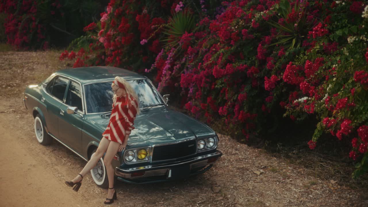 Stylische Frau posiert an einem grünen Oldtimer mit blühenden Blumen