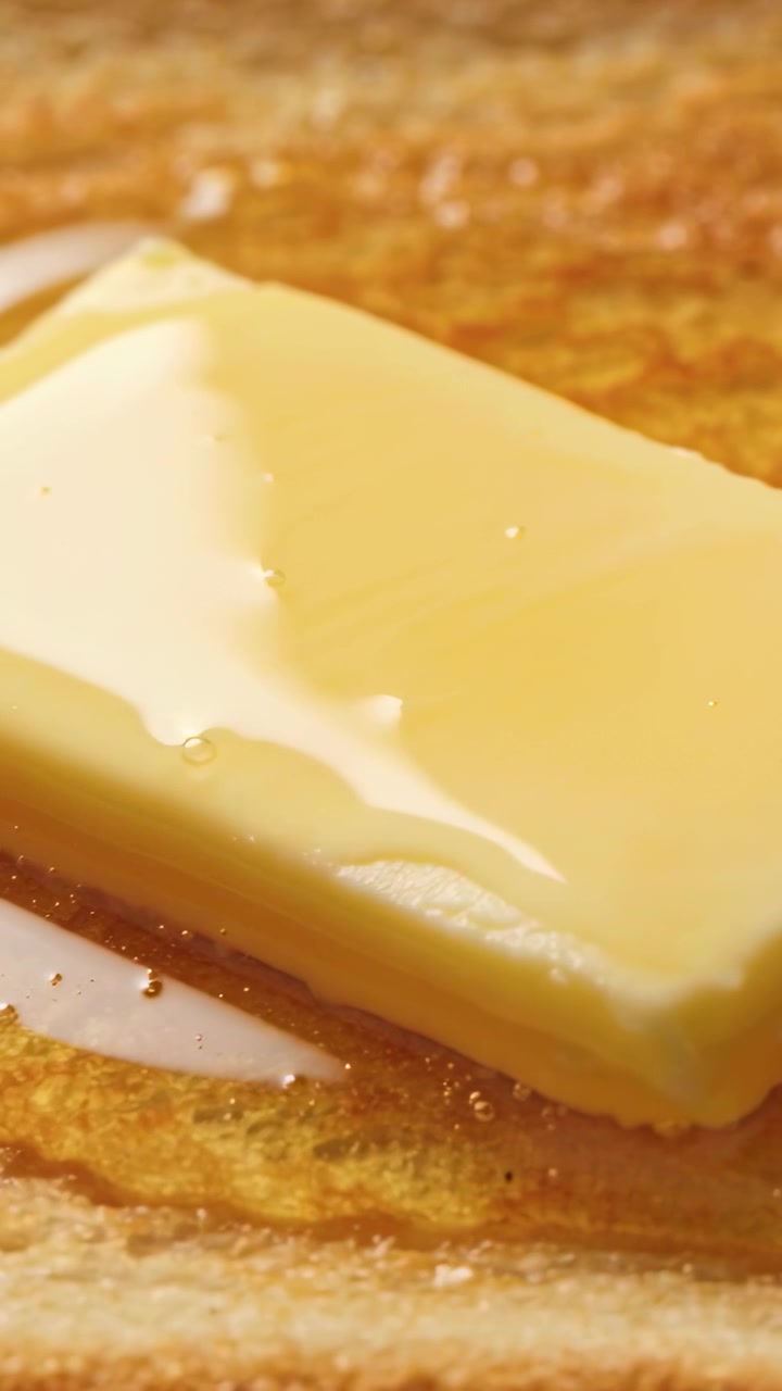 Video vertical: Trozo de mantequilla derritiéndose, ablandándose y formando un charco sobre una tostada caliente, con chorros brillantes empapándose.