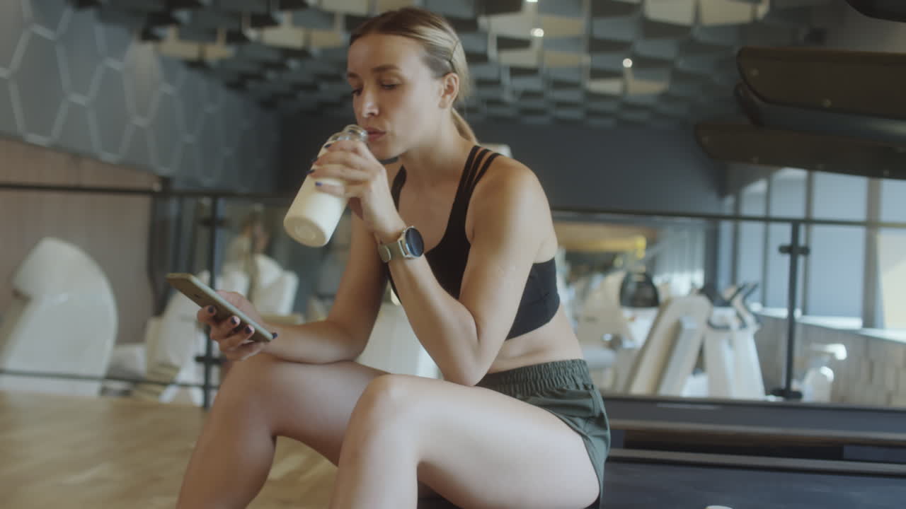 Woman Drinking Water and Texting on Phone in Gym