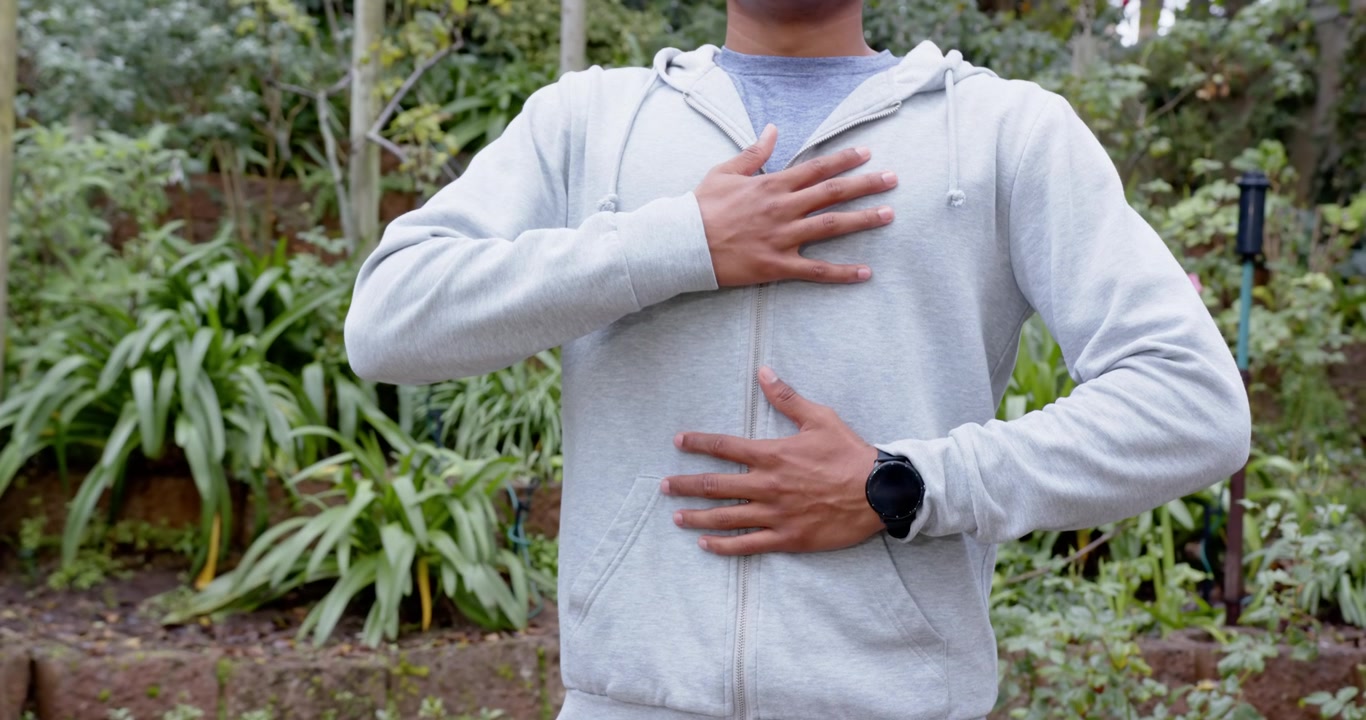 Uomo africano che posiziona le mani sul petto e sull'addome, controllando il respiro in giardino, indossando una felpa con cappuccio