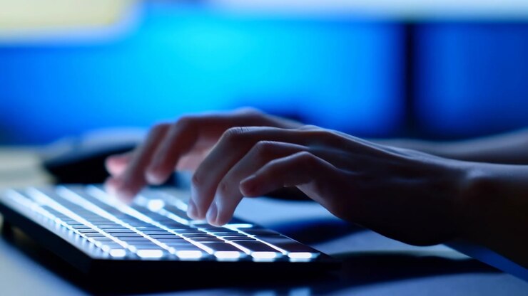 Nahaufnahme von Händen, die auf einer Plastiktastatur tippen, mit einem Computerbildschirm im Hintergrund, die konzentrierte und produktive Büroarbeit in einer modernen und coolen Umgebung mit blauen und schwarzen Tönen zeigt