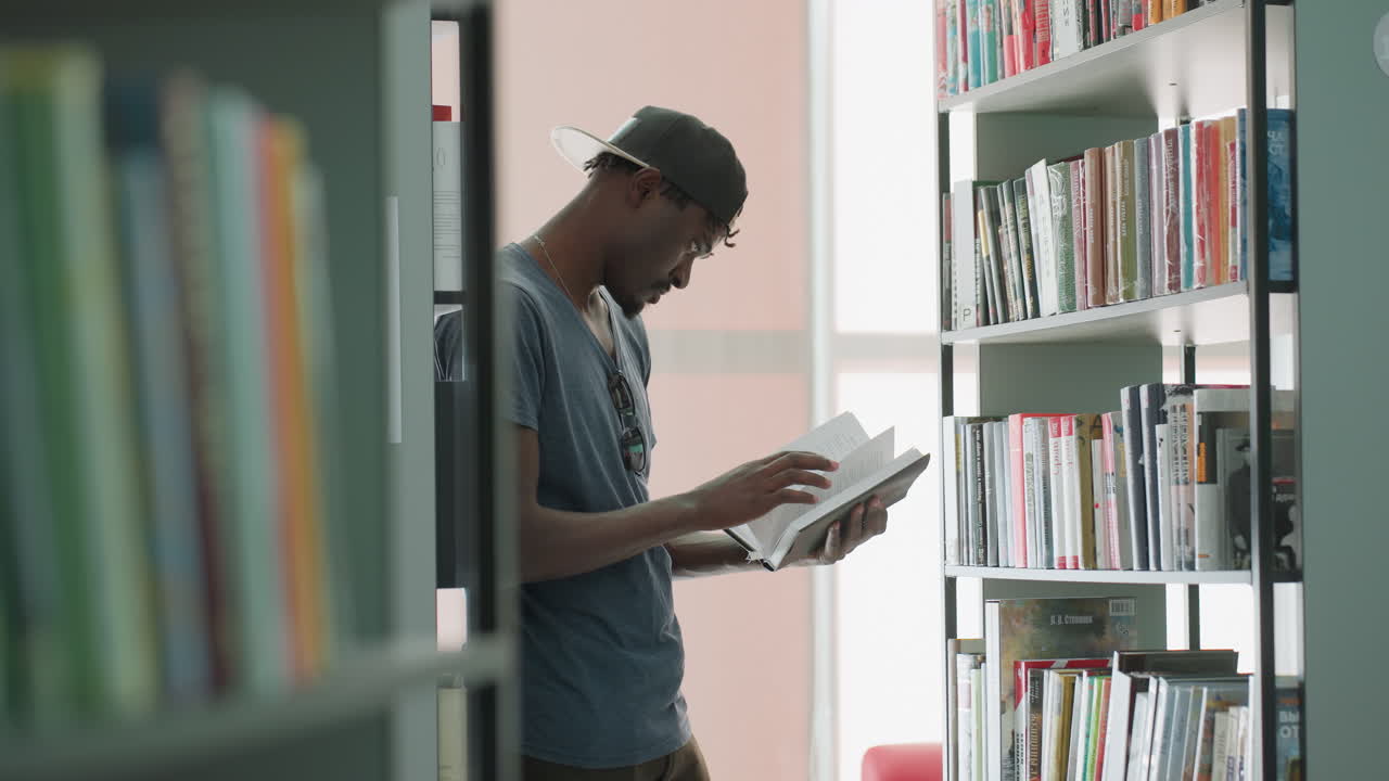 Estudiante de instituto de pie entre estanterías en una luminosa sala de lectura, hojeando pensativamente las páginas de un libro sacado de una pila de la biblioteca, con una postura concentrada cerca de la luz de la ventana