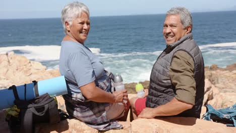 Portrait-of-happy-senior-biracial-couple-in-mountains-resting-at-sea,-in-slow-motion