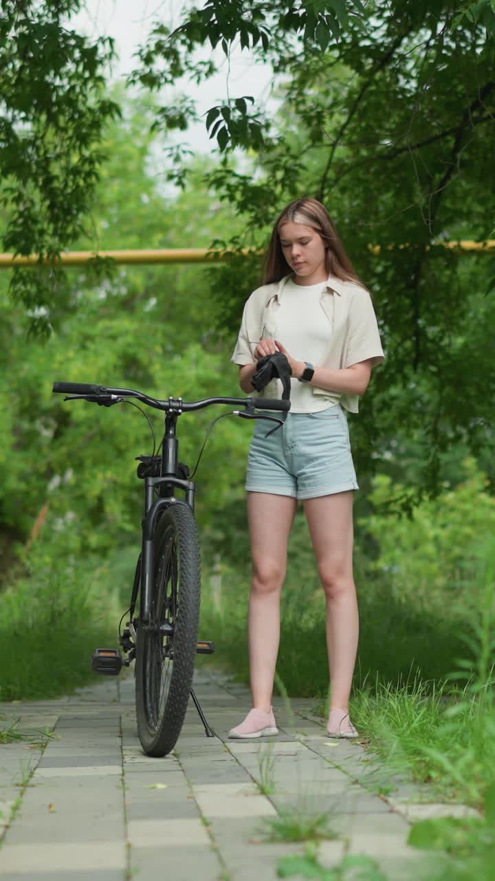 스마트워치가 오른쪽 손에 보이면서 스마트워치를 착용할 준비를 하고 있는 여성이 파크된 자전거에 접근하면서 검은 장갑을 들고 있으며, 배경에는 멀리 지나가는 사람들이 포함되어 있습니다.