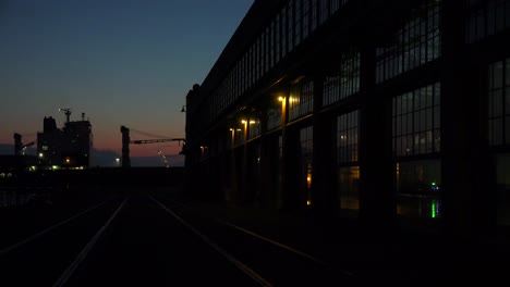 Weitwinkelaufnahme-Eines-Großen-Lagers-Oder-Einer-Fabrik-In-Der-Abenddämmerung-Oder-Bei-Sonnenuntergang,-Während-Ein-Großes-Frachtschiff-In-Der-Ferne-2-.-Vorbeifährt