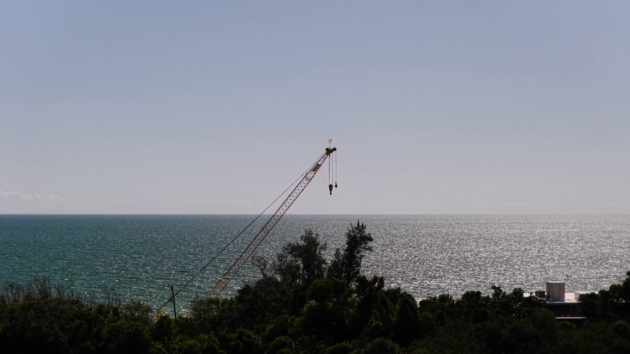 An elevated view of a single lattice boom crane rising above dense trees with the sea and bright sky in the background.