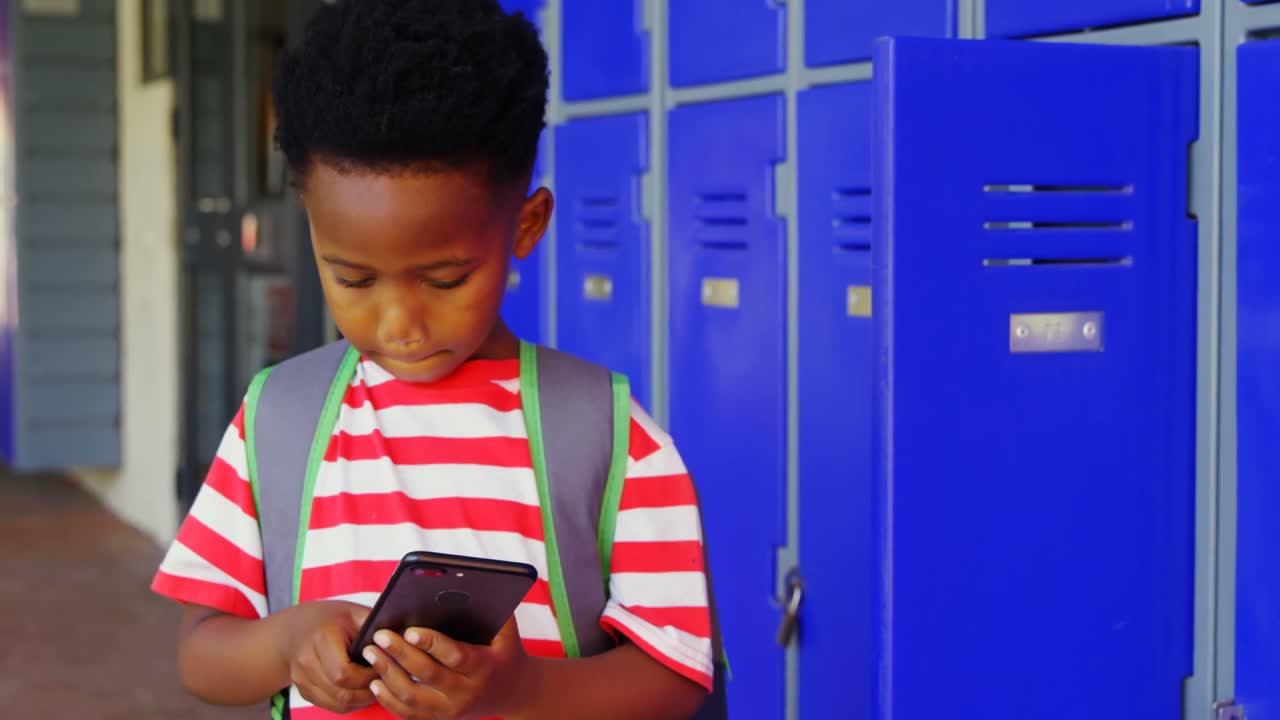 Front view of African American schoolboy with schoolbag using mobile phone in school corridor 4k