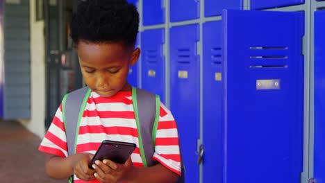 Vista-Frontal-De-Un-Colegial-Afroamericano-Con-Mochila-Usando-Un-Teléfono-Móvil-En-El-Pasillo-De-La-Escuela-4k