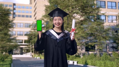asian woman student graduates in cap and gown smiling then showing diploma and green screen smartphone to the camera in front of a magnificent university building