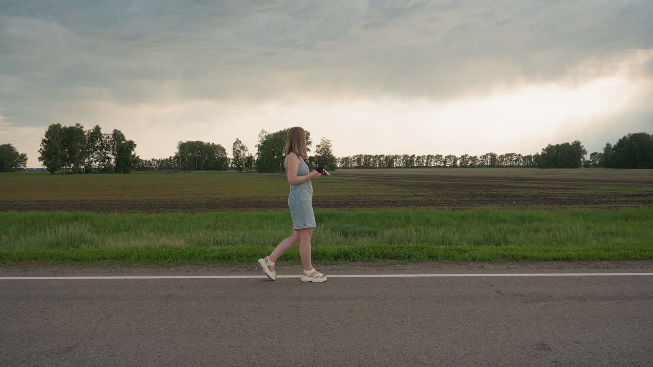 Young woman in denim dress holds colourful pinwheel walking along white road marking beside green field under cloudy summer sky capturing serene motion and playful spirit