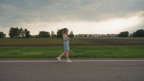 young woman in denim dress holds colourful pinwheel walking along white road marking beside green field under cloudy summer sky capturing serene motion and playful spirit