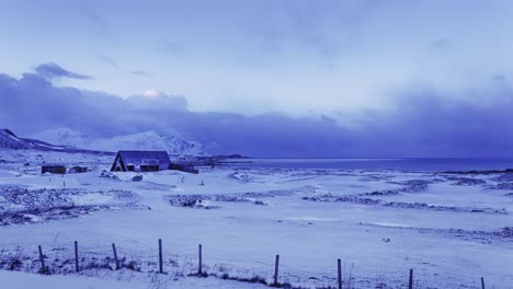 snow covered field by the sea, with fence in the foreground, a building in the middle and mountains in the background