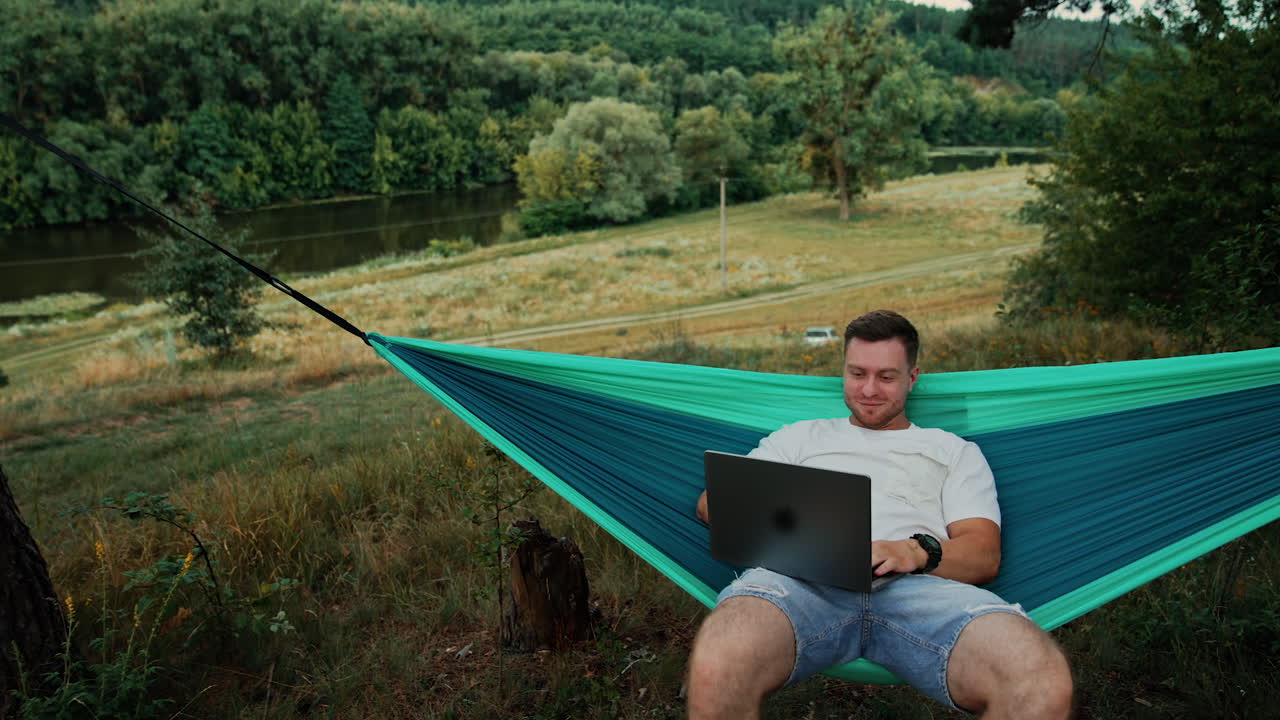 Happy smiling Caucasian man works sitting in the hammock. Man uses laptop swinging in the hammock in nature.