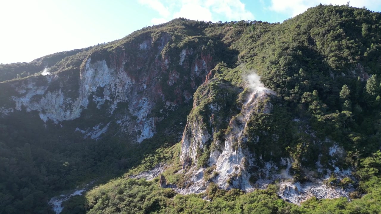 Dolly in con dron hacia una columna de humo que emana de una montaña arcoíris, bosque verde exuberante, Nueva Zelanda