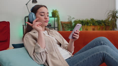 Happy-young-woman-in-wireless-headphones-relaxing-lying-on-sofa-at-home-listening-favorite-music