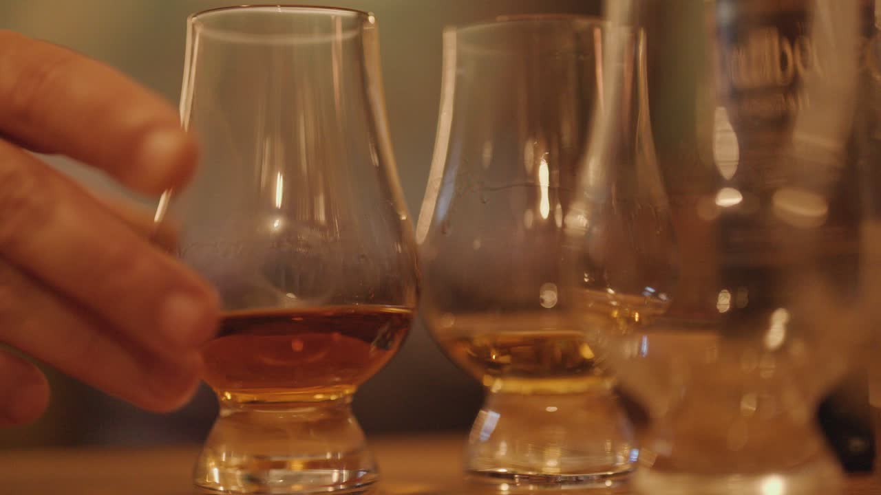 A hand carefully sets a whiskey glass among others on a wooden bar counter in warm, ambient lighting, with a shallow depth of field and steady camera