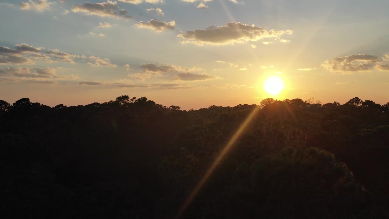 Bright golden sun descending behind the silhouette of a lush forest, casting beautiful sunbeams and lens flare against a sky with scattered clouds, creating a peaceful and serene scene