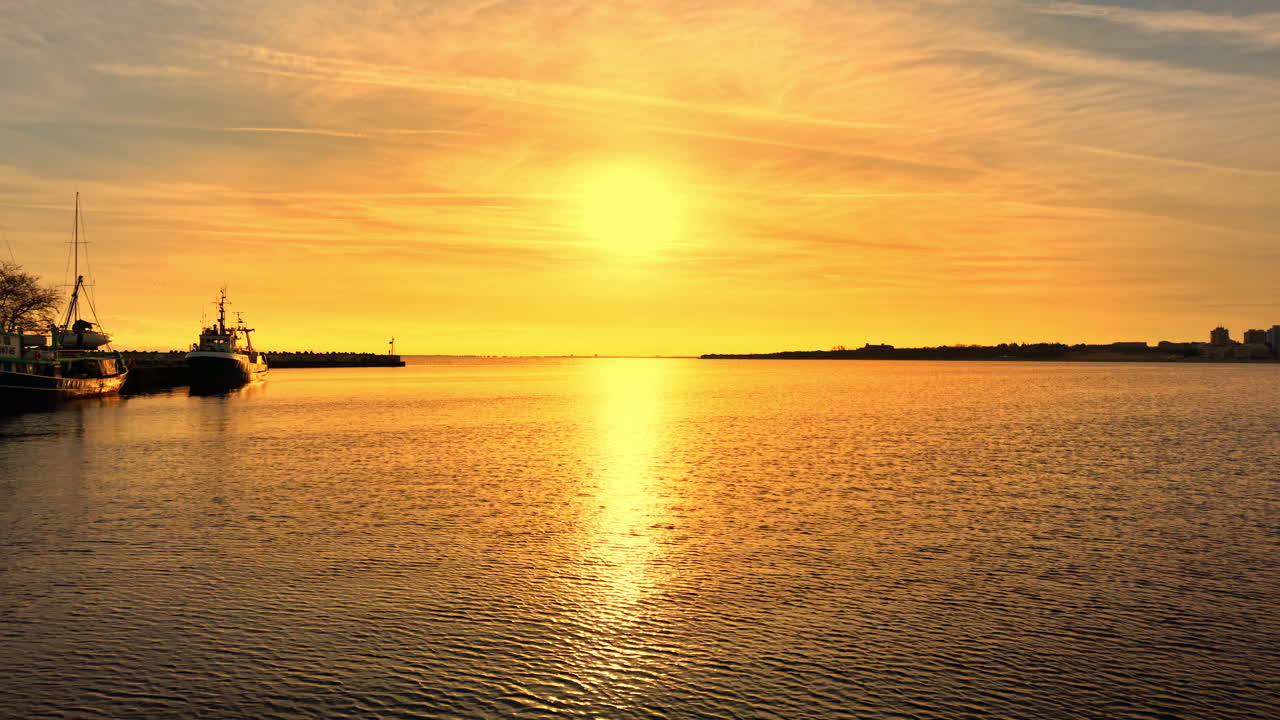 Golden sunset over the sea with fishing boats. Sun rays reflecting on the water surface. Silhouettes of fishing vessels docked at a pier