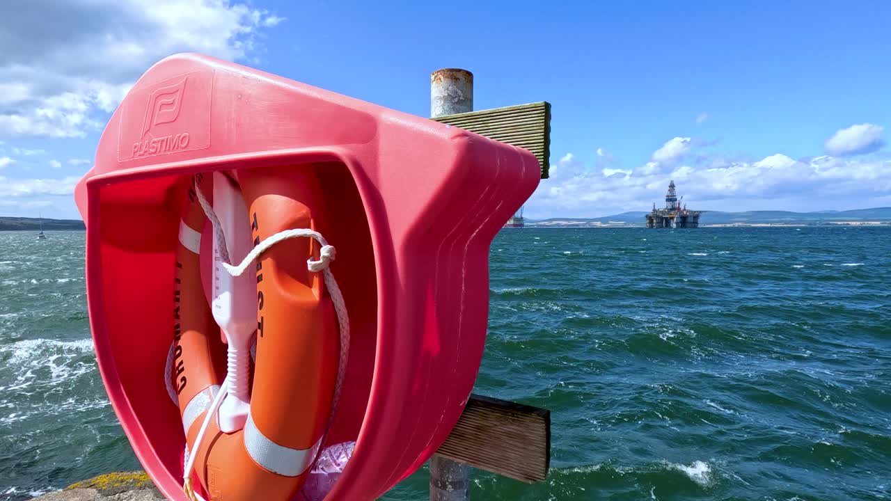 A bright orange lifebuoy ring in a red case is mounted on a wooden pier, with choppy sea and an offshore platform visible under clear daylight. Camera pans slightly